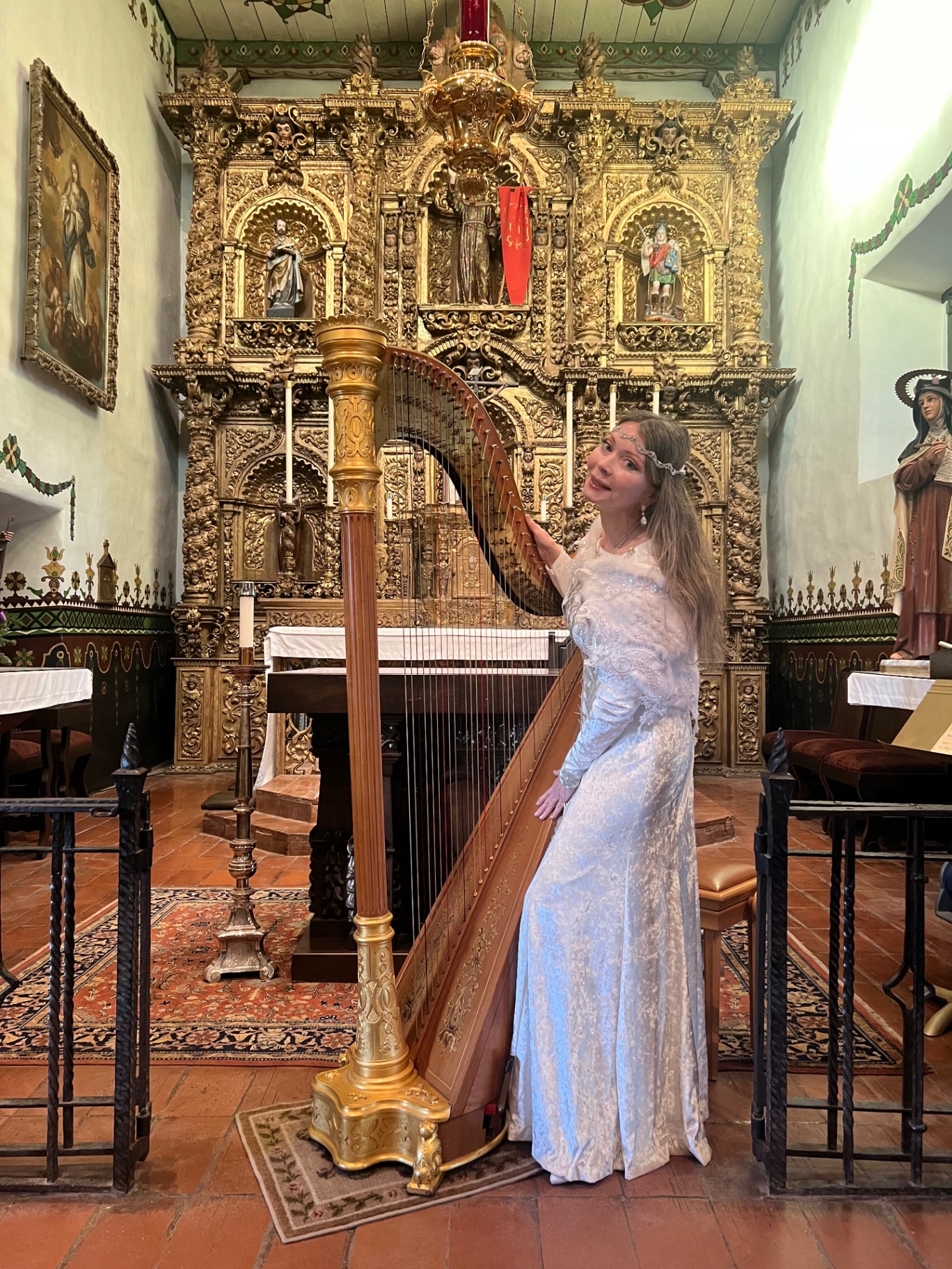 Dr. Marsha Long standing at her concert harp before the gilded altar of a California mission chapel
