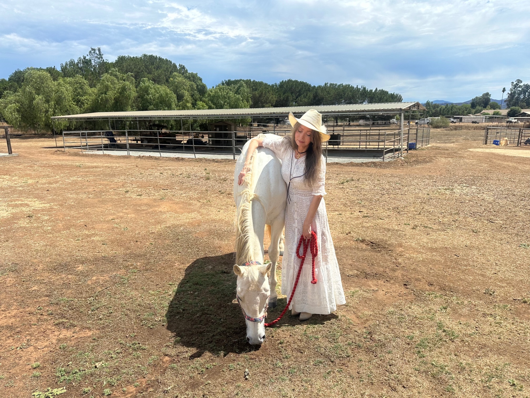 Dr. Marsha Long in a long white lace dress and straw hat standing with her white horse at a San Diego ranch